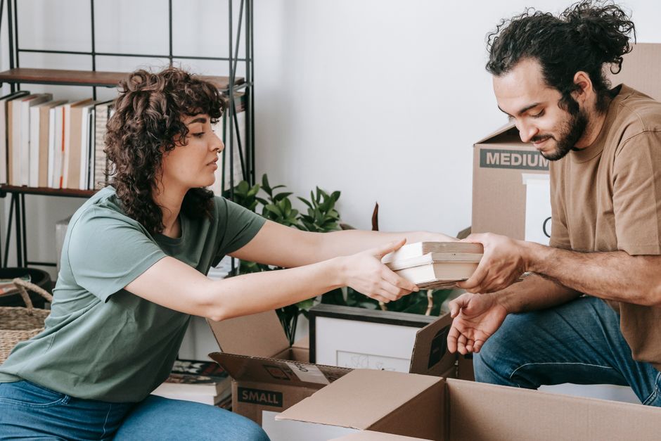 A man with dark, curly hair and a beard is involved in a home relocation process, depicted inside a room with natural lighting. He is wearing a brown T-shirt and is seen carefully lifting or adjusting