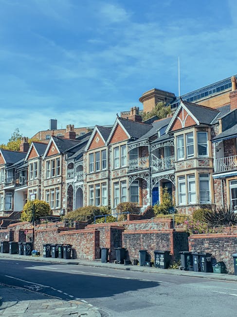 Image showing a row of Victorian-style terraced houses with ornate white balcony railings and decorative brickwork, situated on a street in Chessington. The houses are elevated above the pavement, which is lined with black and green waste bins. A clear blue sky with scattered clouds is visible overhead, and nearby trees provide greenery to the scene. This residential area is part of a typical home relocation setting, where moving companies such as Man with Van Chessington may assist with furniture transport and packing during house removals, with the houses' façades and street environment indicating a high-density urban neighbourhood suitable for relocation services.