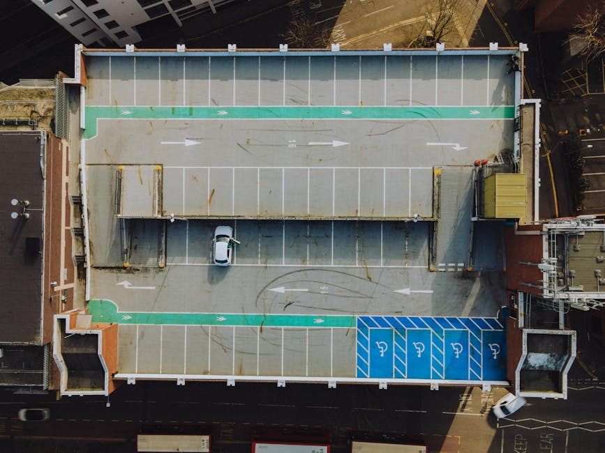 An aerial view of a rooftop parking area on a building, showing designated parking spaces including standard and disabled bays painted in blue with wheelchair symbols. A small white car is parked in one of the accessible spaces, while several unoccupied parking spots remain available. The roof is divided into sections with white parking lines and directional arrows for vehicle movement, along with green markings indicating pedestrian pathways or walking zones. Adjacent to the parking area are parts of the building's structure, such as walls and rooftop equipment, with some surrounding streets visible at the edges of the image. This setting illustrates an urban environment suitable for activities like house removals and furniture transport, with logistics facilitated by companies such as Man with Van Chessington, which specialises in relocations and moving services in the local area.
