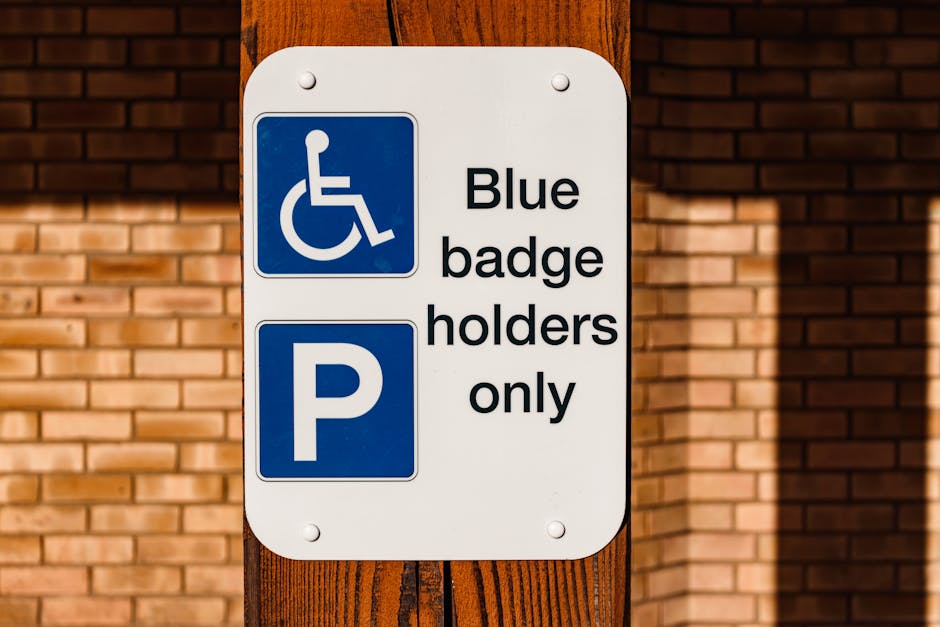 A white rectangular parking permit sign mounted on a vertical wooden post outdoors. The top left corner of the sign features a blue icon of a person in a wheelchair, indicating accessibility, while beneath it, there is large blue parking symbol with a white 'P' in the center. The right side of the sign displays black text reading 'Blue badge holders only'. The background consists of horizontal reddish-brown brickwork with sunlight casting slight shadows, suggesting an exterior setting often encountered during home relocation or furniture transport when parking permits are relevant. The sign's placement on the wooden post indicates a designated parking area for disabled badge holders, which Man with Van Chessington may need to consider when planning house removals or loading and unloading furniture. The overall scene reflects the necessary logistical considerations for moving services in environments requiring accessible parking arrangements within urban or residential areas.