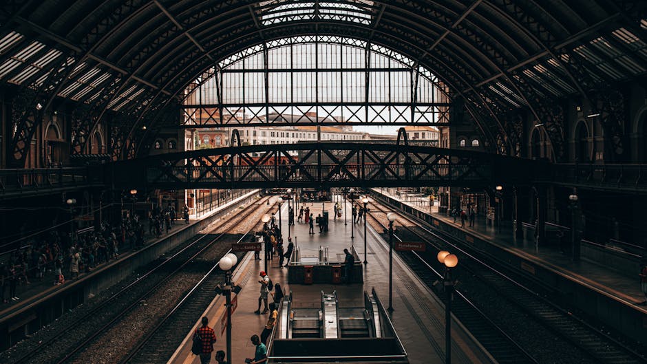 The image depicts a large, historic train station with a high, arched metal and glass roof allowing natural light to illuminate the interior. Inside, multiple railway tracks run parallel along the platform area, where several travelers are waiting or walking. A few individuals are visible carrying or loading luggage, while some are standing near the edges of the platform. The station features vintage-style lighting fixtures and signage, with an open, spacious environment conducive to home relocation and furniture transport needs. The station's architecture and busy atmosphere reflect the typical scene during a moving or packing and moving process, with people arriving or departing, and the station serving as a location for station-to-station or local transport, supported by the professional removals services of Man with Van Chessington.
