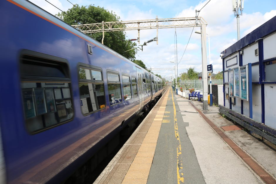 The image depicts a large, historic train station with a high, arched metal and glass roof allowing natural light to illuminate the interior. Inside, multiple railway tracks run parallel along the platform area, where several travelers are waiting or walking. A few individuals are visible carrying or loading luggage, while some are standing near the edges of the platform. The station features vintage-style lighting fixtures and signage, with an open, spacious environment conducive to home relocation and furniture transport needs. The station's architecture and busy atmosphere reflect the typical scene during a moving or packing and moving process, with people arriving or departing, and the station serving as a location for station-to-station or local transport, supported by the professional removals services of Man with Van Chessington.