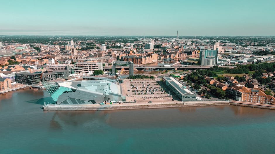 An aerial view of the Royal Borough of Kingston in Chessington, showing a mix of modern and traditional buildings along the riverbank, with a large, contemporary cultural or commercial structure featuring angular, metallic surfaces in the foreground. The surrounding area includes residential housing, parking lots, and roads, with a landscape of trees and greenery in the background. In the image, a man from Man with Van Chessington is engaged in a home relocation process, loading or unloading furniture and cardboard boxes from a van parked near the riverside, with some items covered in protective plastic wrap and moving blankets. The scene is captured in daylight with clear visibility, highlighting the logistics involved in packing and furniture transport as part of a move aligned with house removals and relocation services.