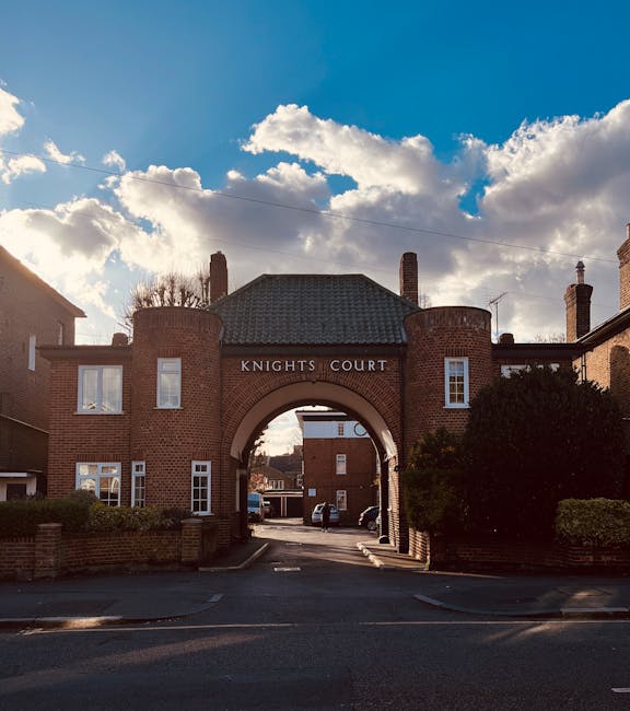 The image shows the entrance archway of Knights Court, a residential building in Chessington with a brick façade, two rounded towers on either side, and a central archway leading into a parking area. The building has multiple windows, some with white frames, and a paved driveway beneath the arch. The sky above is partly cloudy with sunlight illuminating parts of the structure, casting shadows on the pavement. To the right of the entrance, dense green bushes and small trees are visible, contributing to the estate's appearance. No moving activity or furniture is visible in this image, but the setting suggests a suburban area typical for house removals or relocation services. Occasionally, Man with Van Chessington operates in this area to facilitate home relocations and furniture transport, especially in properties like Knights Court.
