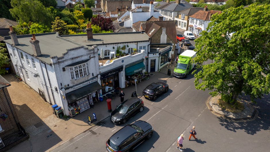 An aerial view of a busy street scene on Chessington High Street, featuring a small white building with a flat roof, accommodating a shop with an awning and display windows. Several parked vehicles, i
