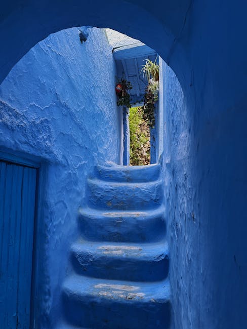 A narrow, curved stone staircase inside a blue-painted building leading upward towards an open doorway, with potted plants and a blue shutter visible through the doorway. The blue walls and steps are textured with visible paint strokes and wear, creating a vibrant, picturesque setting typical of Mediterranean or coastal house exteriors. Natural light illuminates the scene through the doorway, highlighting the contours of the steps and the bright scene beyond. The setting appears to be part of a home or a domestic interior, possibly used during a house relocation or packing and moving process by Man with Van Chessington, with the space designed for easy access and movement of household items between rooms or to an outdoor area.