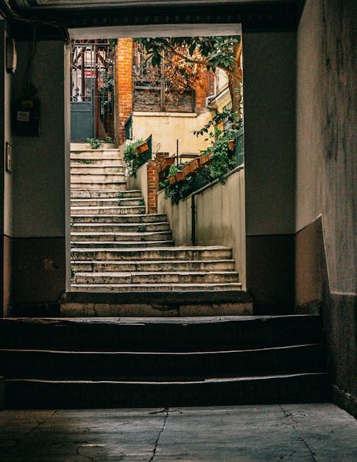 A view through a dark, indoor stairwell showing the outdoor area beyond. The outdoor space features a set of ascending concrete stairs with visible wear and slight unevenness, leading up to a small landing with additional steps. To the right, a cream-colored wall borders a garden area with green foliage and potted plants, some of which are placed on a small ledge. To the left, brick stairs and a black metal railing are partially visible, leading further up to a brick building with a window and a wooden fence. The scene is illuminated by natural daylight, highlighting the textured surfaces of the stairs and walls. This image captures the typical environment and architectural features associated with home relocation and moving logistics, as part of a professional removal service such as Man with Van Chessington preparing for furniture transport and packing through narrow staircases and restricted doorways.