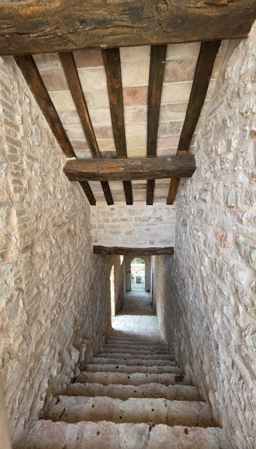 A narrow, curved stone staircase inside a blue-painted building leading upward towards an open doorway, with potted plants and a blue shutter visible through the doorway. The blue walls and steps are textured with visible paint strokes and wear, creating a vibrant, picturesque setting typical of Mediterranean or coastal house exteriors. Natural light illuminates the scene through the doorway, highlighting the contours of the steps and the bright scene beyond. The setting appears to be part of a home or a domestic interior, possibly used during a house relocation or packing and moving process by Man with Van Chessington, with the space designed for easy access and movement of household items between rooms or to an outdoor area.