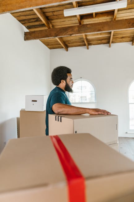 A man with dark curly hair and a beard, wearing a blue t-shirt, is standing inside a room with a wooden-beamed ceiling and white walls. He is positioned behind several cardboard boxes, some of which are sealed with red packing tape, and is resting his arms on top of one of the boxes, preparing for the home relocation process. The room has natural light coming through two arched windows, and the floor appears to be light-colored wood or laminate. The cardboard boxes vary in size and are stacked or arranged in a way that suggests they are being prepared for transport or moving. The overall scene captures the loading and packing phase of furniture transport, related to house removals services provided by Man with Van Chessington, with an emphasis on careful handling and organized packing as part of a last-minute or urgent move.