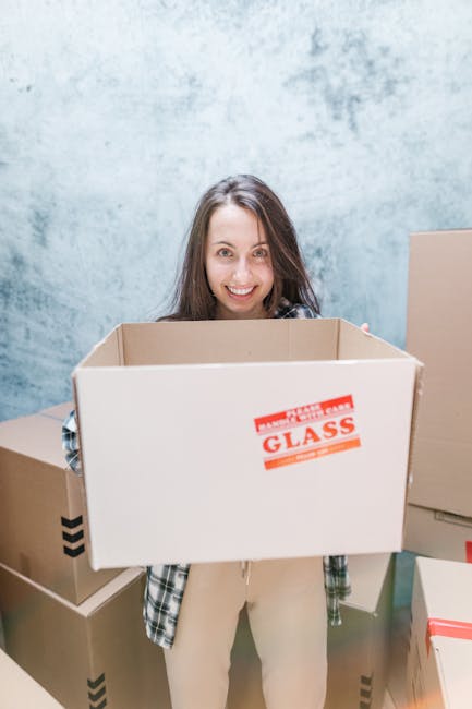 A woman with long brown hair smiling while holding an empty brown cardboard moving box labeled 'GLASS' in red print, standing in a room with light blue textured walls. Several other cardboard boxes, some sealed with packing tape and others open, are stacked around her. The boxes vary in size, with some covered in black tape or red tape, indicating packing and moving preparations. The scene captures the interior of a property during the packing and loading process, suggesting a home relocation. Man with Van Chessington, a professional removals service, is implied to be involved in the furniture transport and packing logistics depicted in the image.
