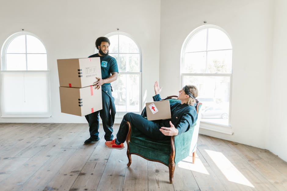 A man with dark curly hair and a beard, wearing a blue t-shirt, is standing inside a room with a wooden-beamed ceiling and white walls. He is positioned behind several cardboard boxes, some of which are sealed with red packing tape, and is resting his arms on top of one of the boxes, preparing for the home relocation process. The room has natural light coming through two arched windows, and the floor appears to be light-colored wood or laminate. The cardboard boxes vary in size and are stacked or arranged in a way that suggests they are being prepared for transport or moving. The overall scene captures the loading and packing phase of furniture transport, related to house removals services provided by Man with Van Chessington, with an emphasis on careful handling and organized packing as part of a last-minute or urgent move.